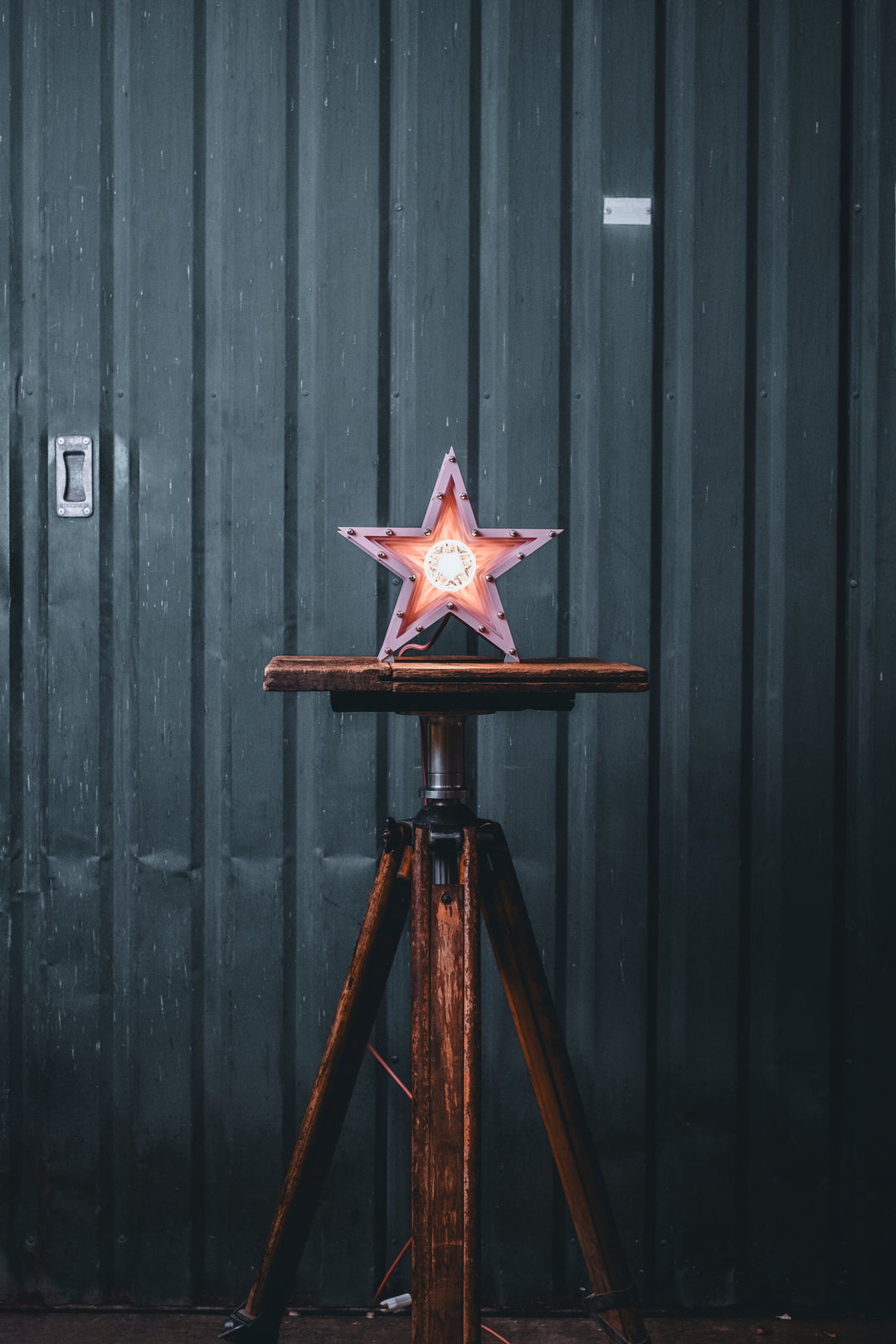 Handcrafted pink star-shaped lamp with layered metal fins and exposed central bulb, sitting on a rustic wooden table.