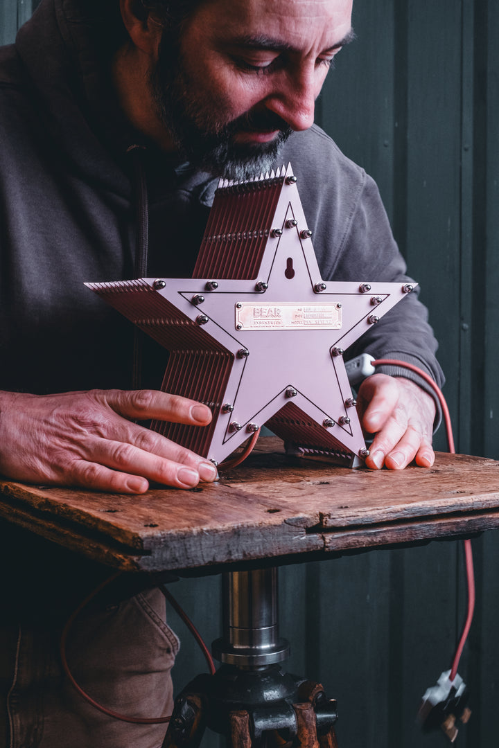 Handcrafted pink star-shaped lamp with layered metal fins and exposed central bulb, sitting on a rustic wooden table.