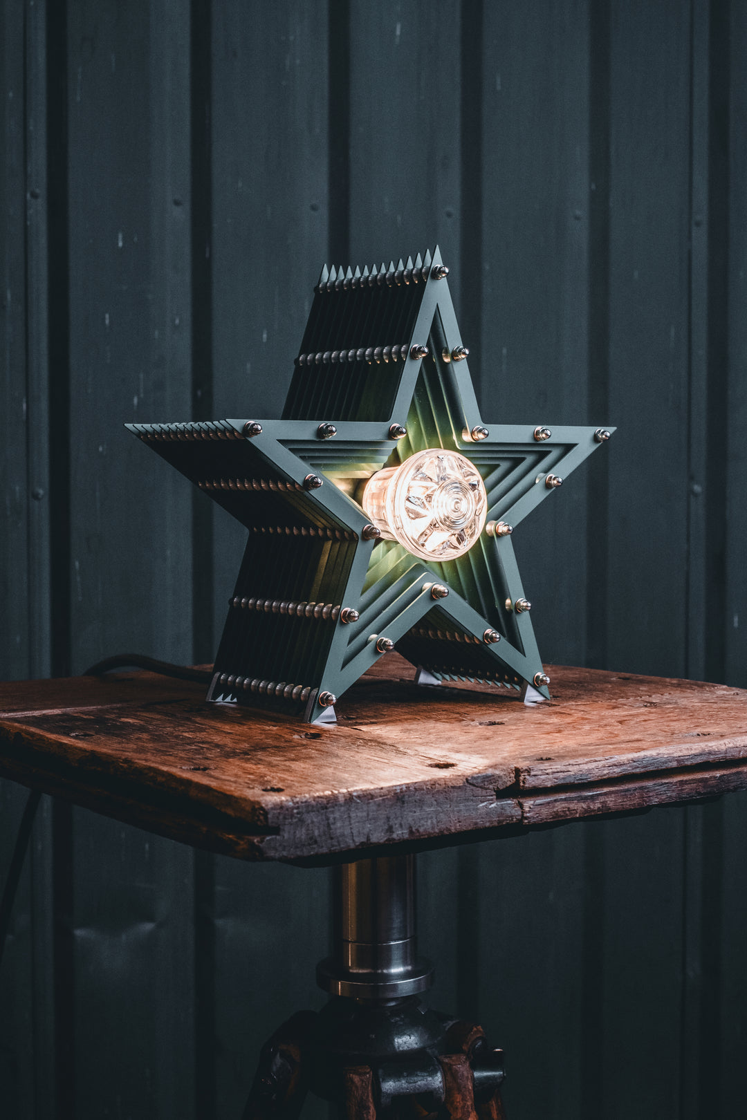 Deep green handcrafted star-shaped lamp with a central illuminated bulb, constructed from layered aluminium with stainless steel fixings and precision-machined spacers. Displayed on a rustic wood and metal tripod table.