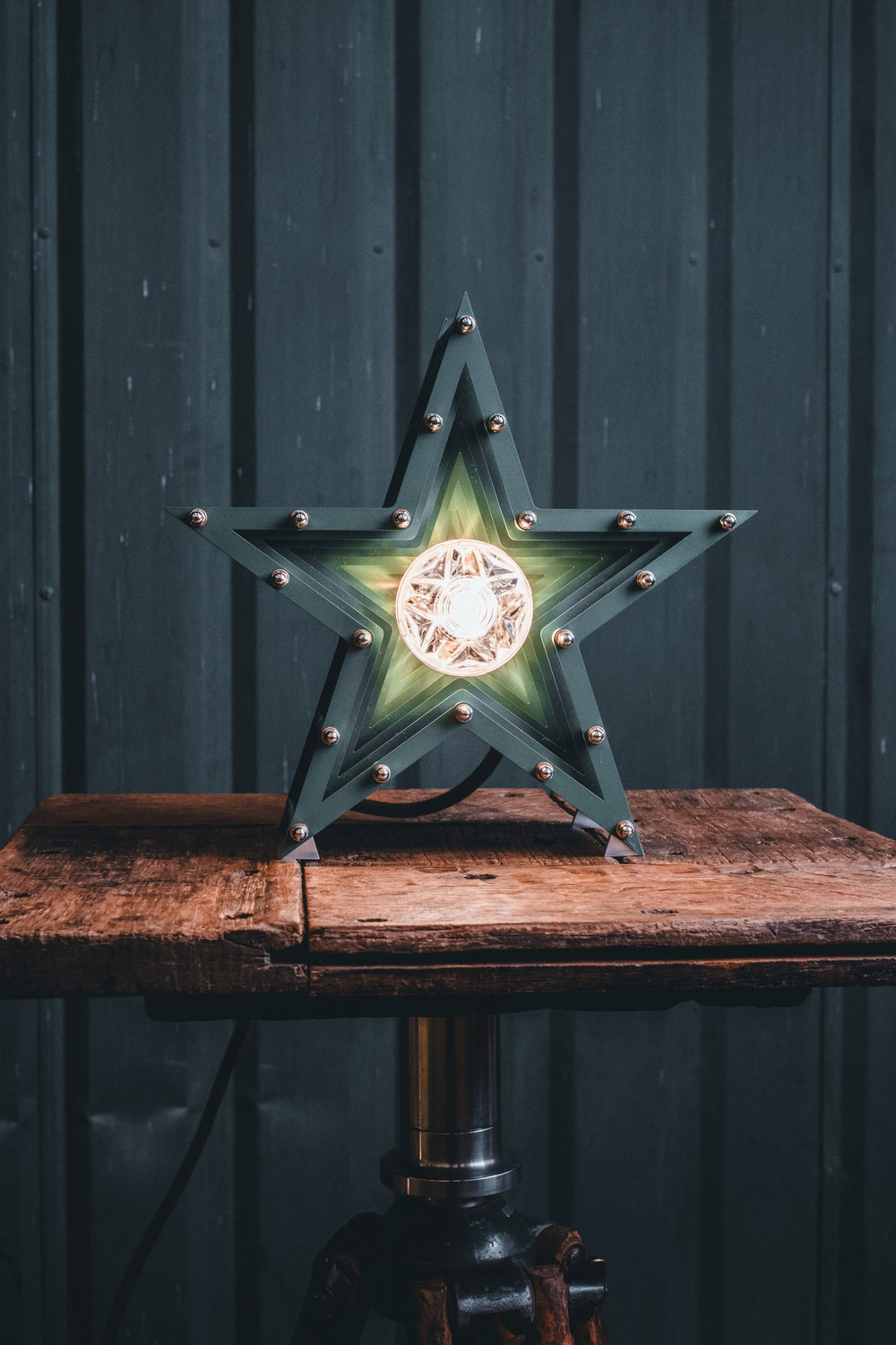 Deep green handcrafted star-shaped lamp with a central illuminated bulb, constructed from layered aluminium with stainless steel fixings and precision-machined spacers. Displayed on a rustic wood and metal tripod table.