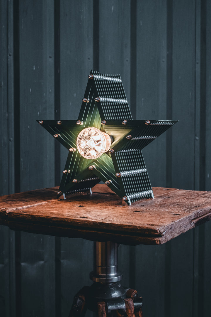 Deep green handcrafted star-shaped lamp with a central illuminated bulb, constructed from layered aluminium with stainless steel fixings and precision-machined spacers. Displayed on a rustic wood and metal tripod table.