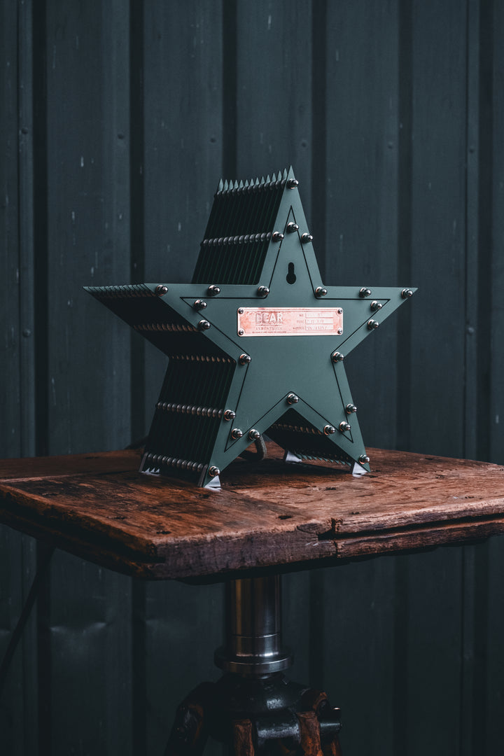 Deep green handcrafted star-shaped lamp with a central illuminated bulb, constructed from layered aluminium with stainless steel fixings and precision-machined spacers. Displayed on a rustic wood and metal tripod table.