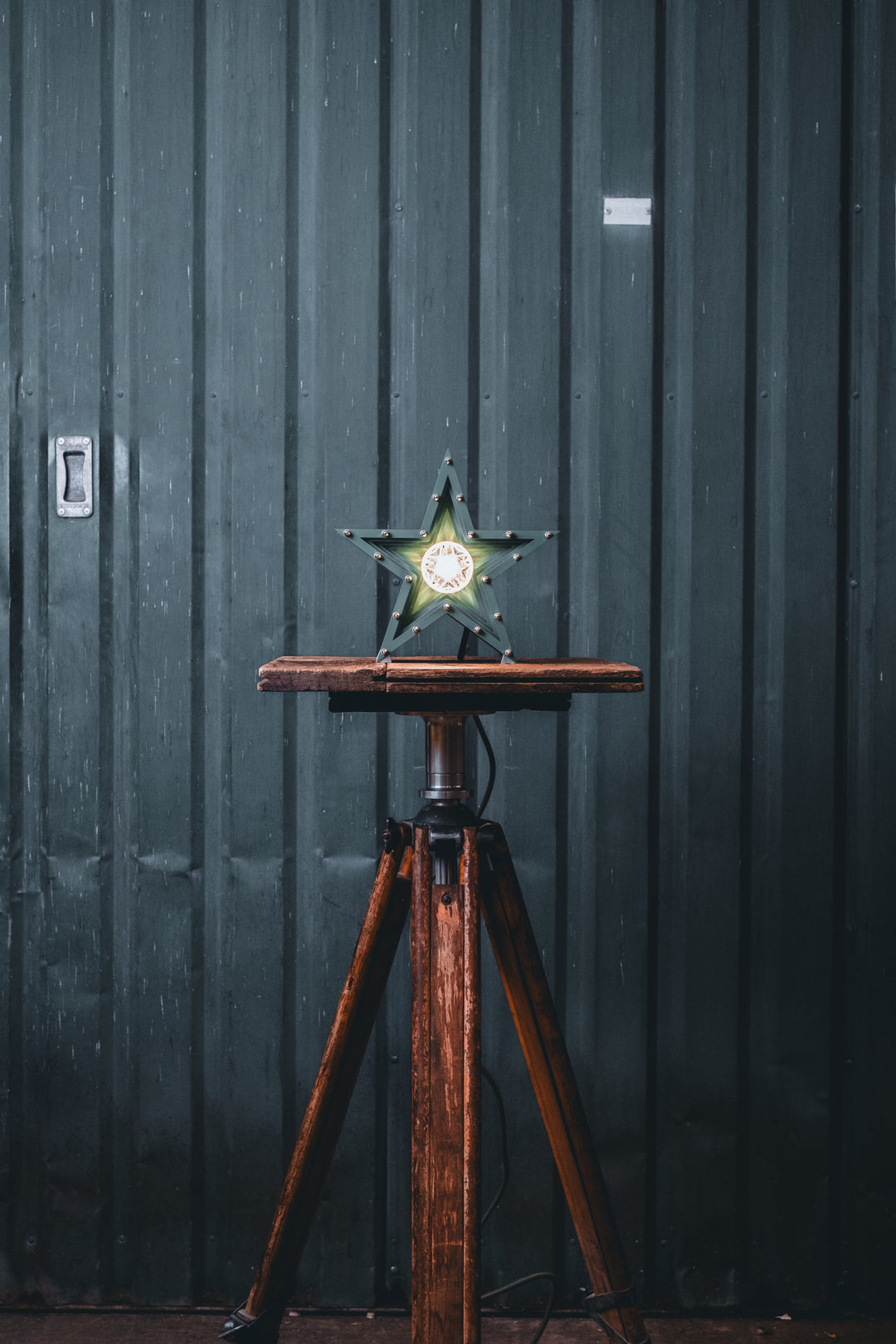 Deep green handcrafted star-shaped lamp with a central illuminated bulb, constructed from layered aluminium with stainless steel fixings and precision-machined spacers. Displayed on a rustic wood and metal tripod table.