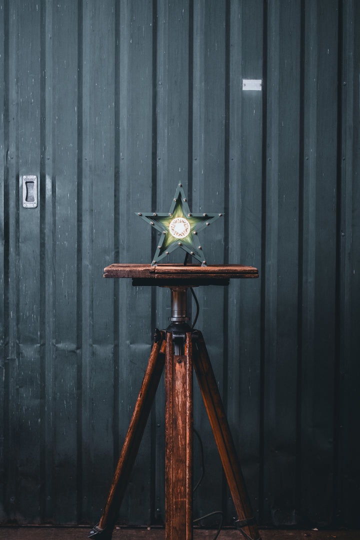 Deep green handcrafted star-shaped lamp with a central illuminated bulb, constructed from layered aluminium with stainless steel fixings and precision-machined spacers. Displayed on a rustic wood and metal tripod table.