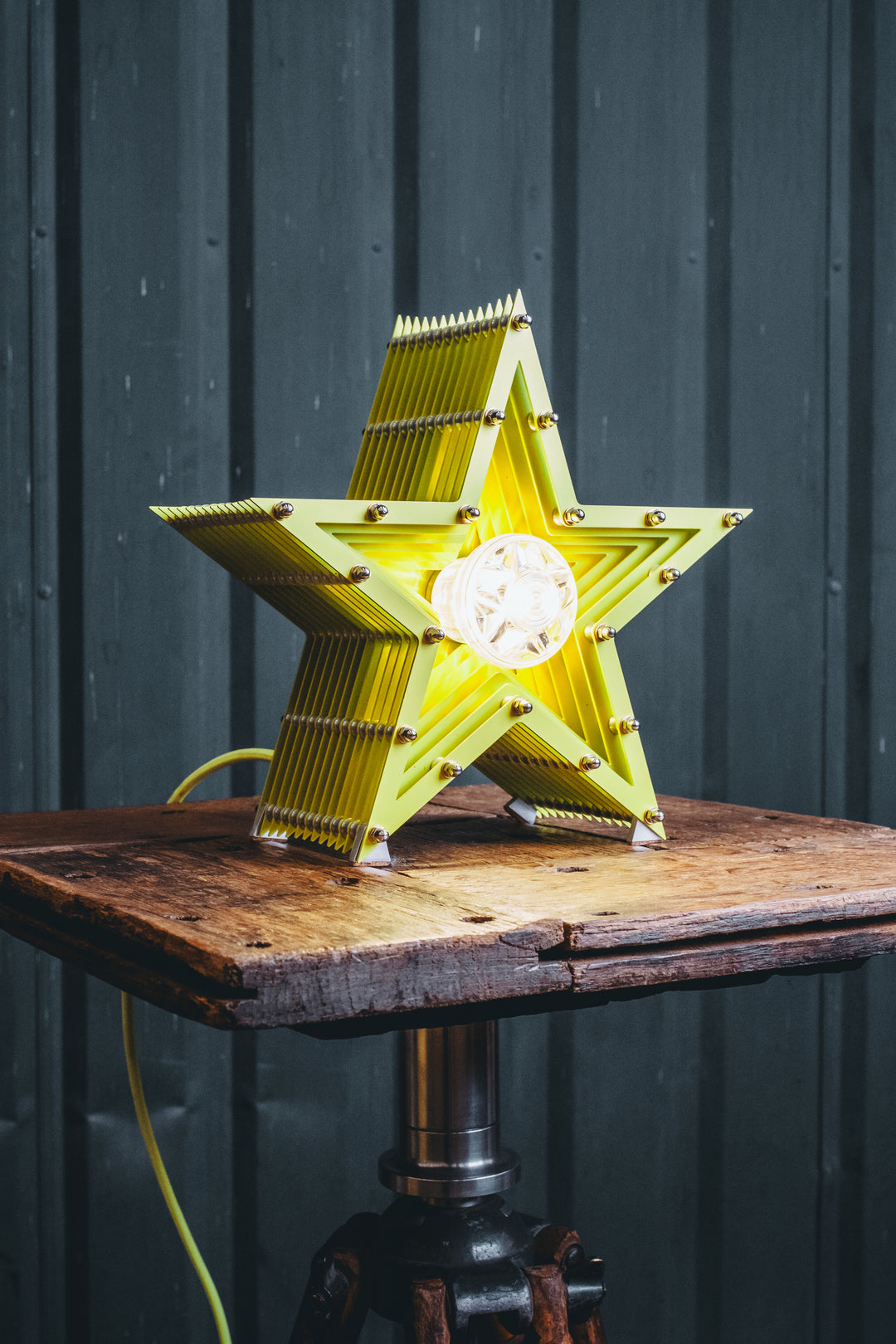 Handcrafted star-shaped lamp in vivid chartreuse yellow, constructed from layered aluminium with precision spacers and a central bulb encased in a bespoke glass surround. Displayed on a rustic wooden table against a dark vertical panel background.