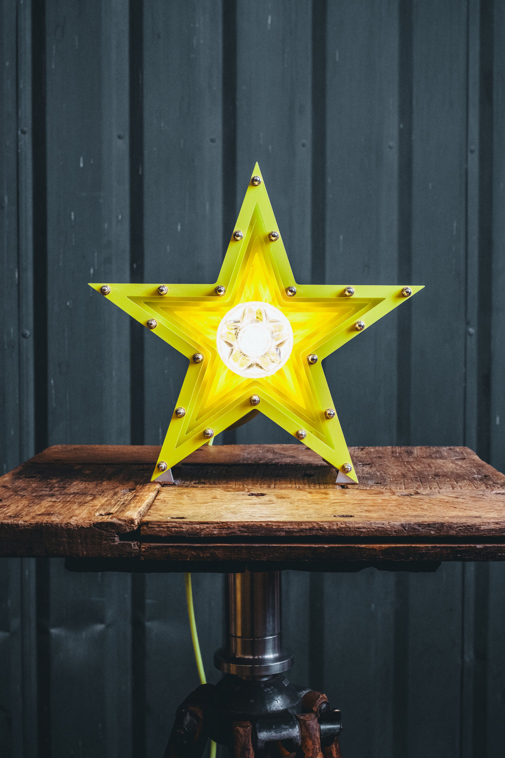 Handcrafted star-shaped lamp in vivid chartreuse yellow, constructed from layered aluminium with precision spacers and a central bulb encased in a bespoke glass surround. Displayed on a rustic wooden table against a dark vertical panel background.