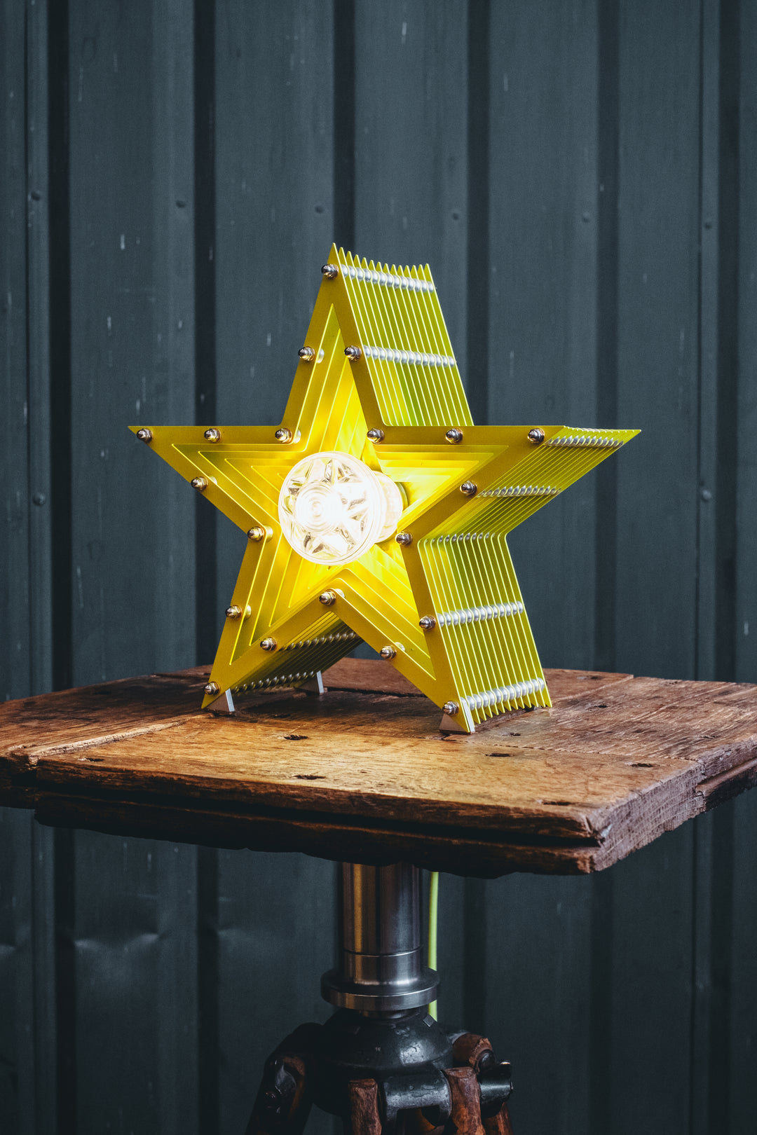 Handcrafted star-shaped lamp in vivid chartreuse yellow, constructed from layered aluminium with precision spacers and a central bulb encased in a bespoke glass surround. Displayed on a rustic wooden table against a dark vertical panel background.