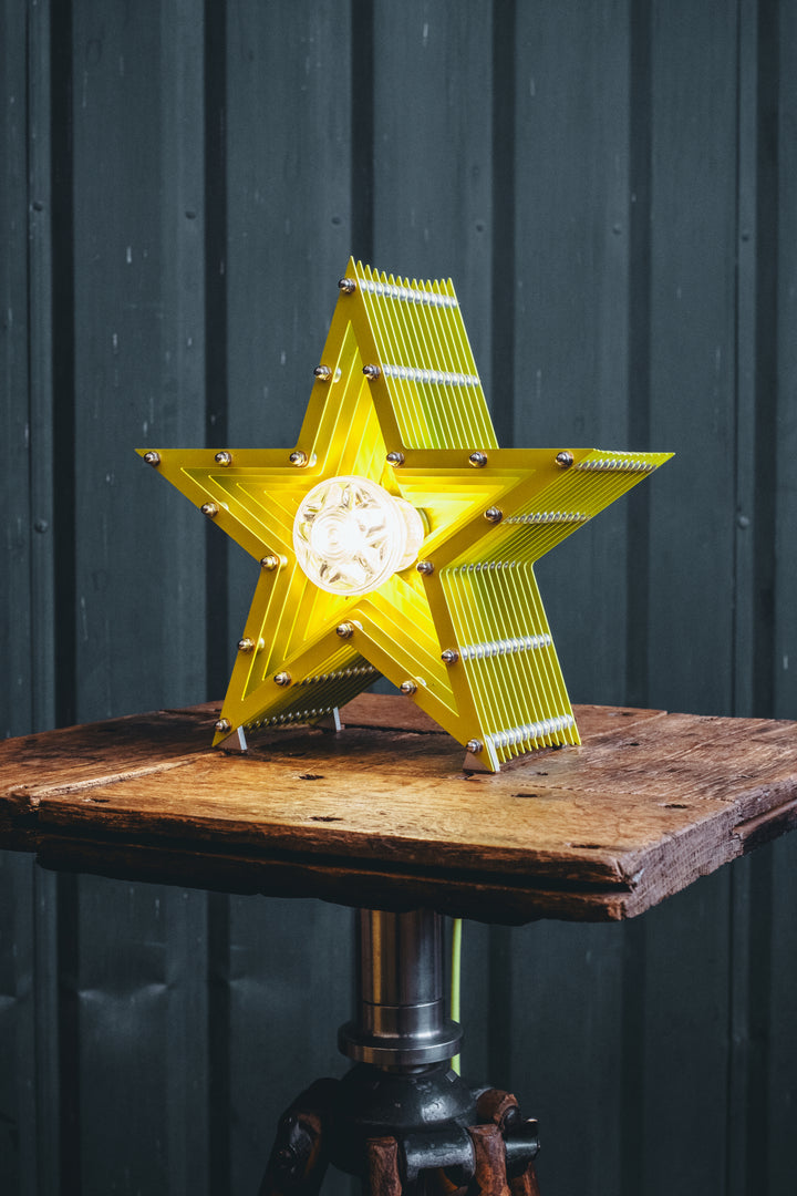 Handcrafted star-shaped lamp in vivid chartreuse yellow, constructed from layered aluminium with precision spacers and a central bulb encased in a bespoke glass surround. Displayed on a rustic wooden table against a dark vertical panel background.