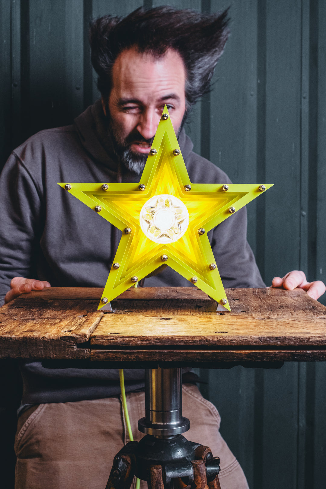 Handcrafted star-shaped lamp in vivid chartreuse yellow, constructed from layered aluminium with precision spacers and a central bulb encased in a bespoke glass surround. Displayed on a rustic wooden table against a dark vertical panel background.