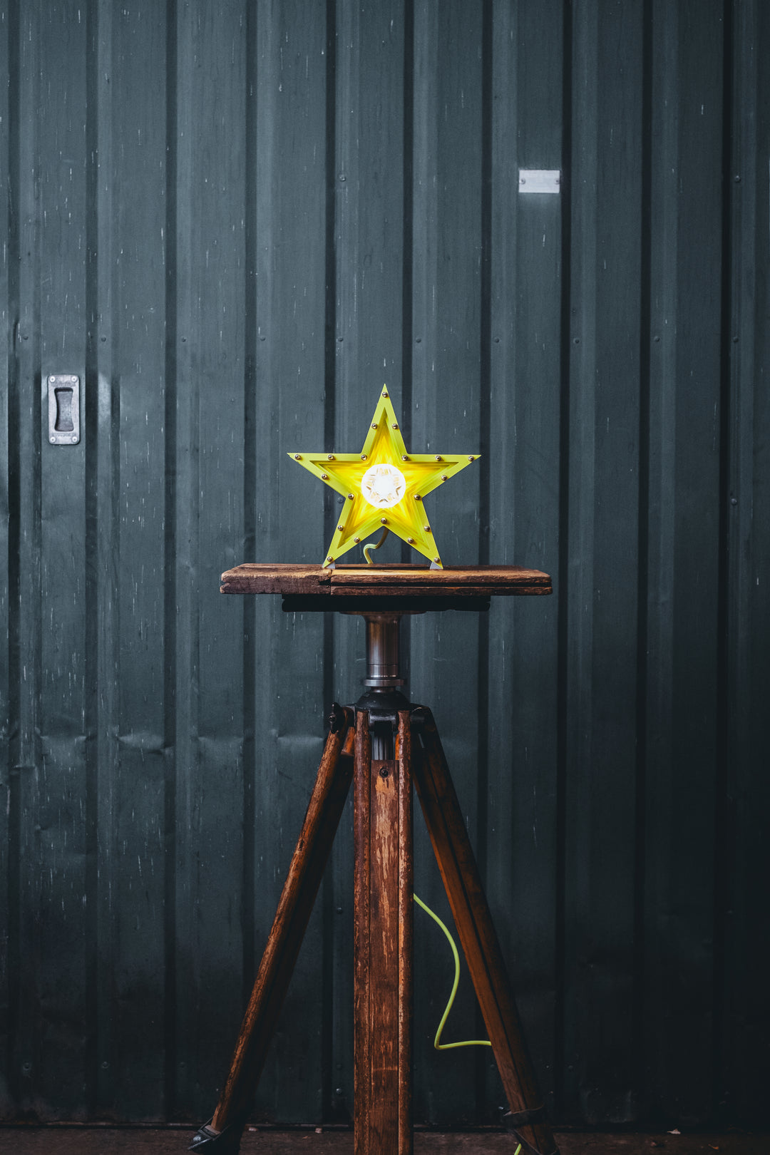 Handcrafted star-shaped lamp in vivid chartreuse yellow, constructed from layered aluminium with precision spacers and a central bulb encased in a bespoke glass surround. Displayed on a rustic wooden table against a dark vertical panel background.
