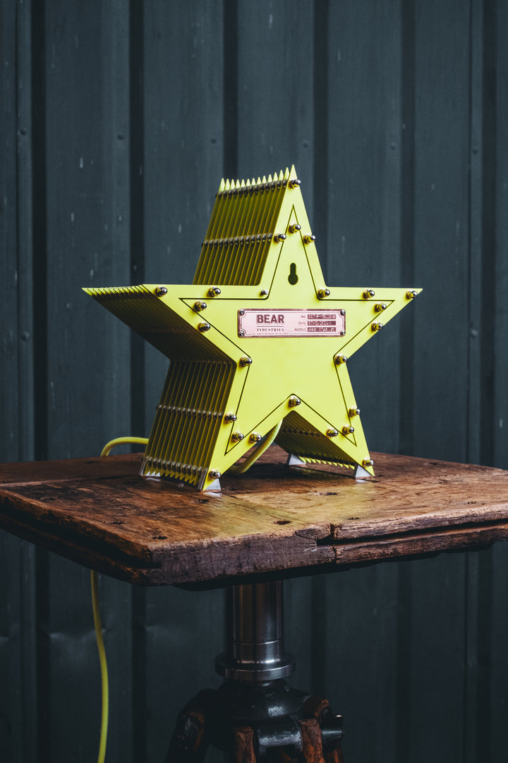 Handcrafted star-shaped lamp in vivid chartreuse yellow, constructed from layered aluminium with precision spacers and a central bulb encased in a bespoke glass surround. Displayed on a rustic wooden table against a dark vertical panel background.