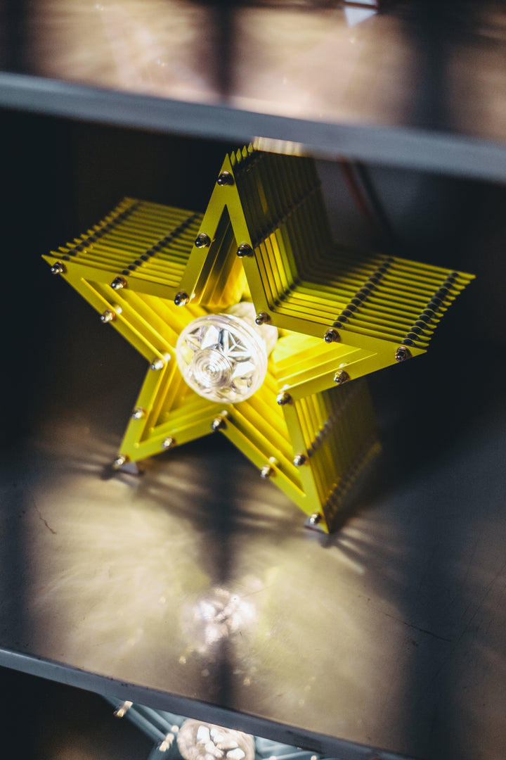 Handcrafted star-shaped lamp in vivid chartreuse yellow, constructed from layered aluminium with precision spacers and a central bulb encased in a bespoke glass surround. Displayed on a rustic wooden table against a dark vertical panel background.