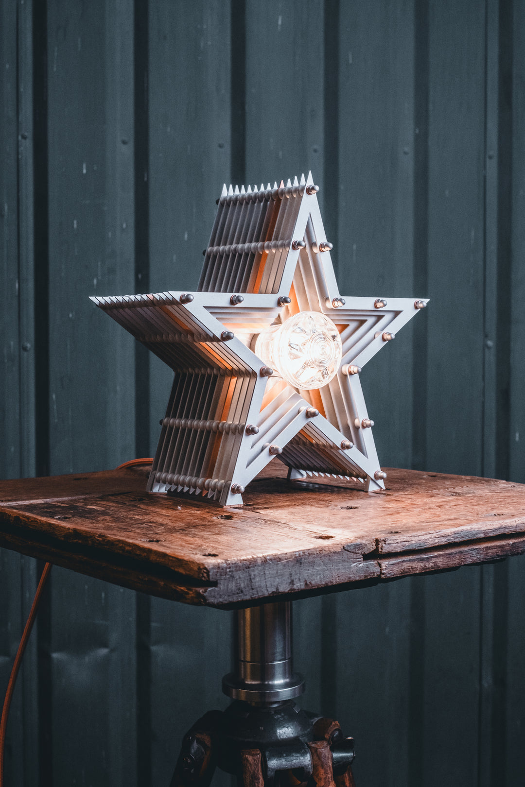 Side view of a Mini Fairground Star lamp in brushed aluminium finish, featuring layered star construction with exposed studding and central illuminated bulb on a rustic wooden table.