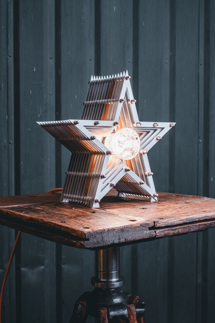 Side view of a Mini Fairground Star lamp in brushed aluminium finish, featuring layered star construction with exposed studding and central illuminated bulb on a rustic wooden table.