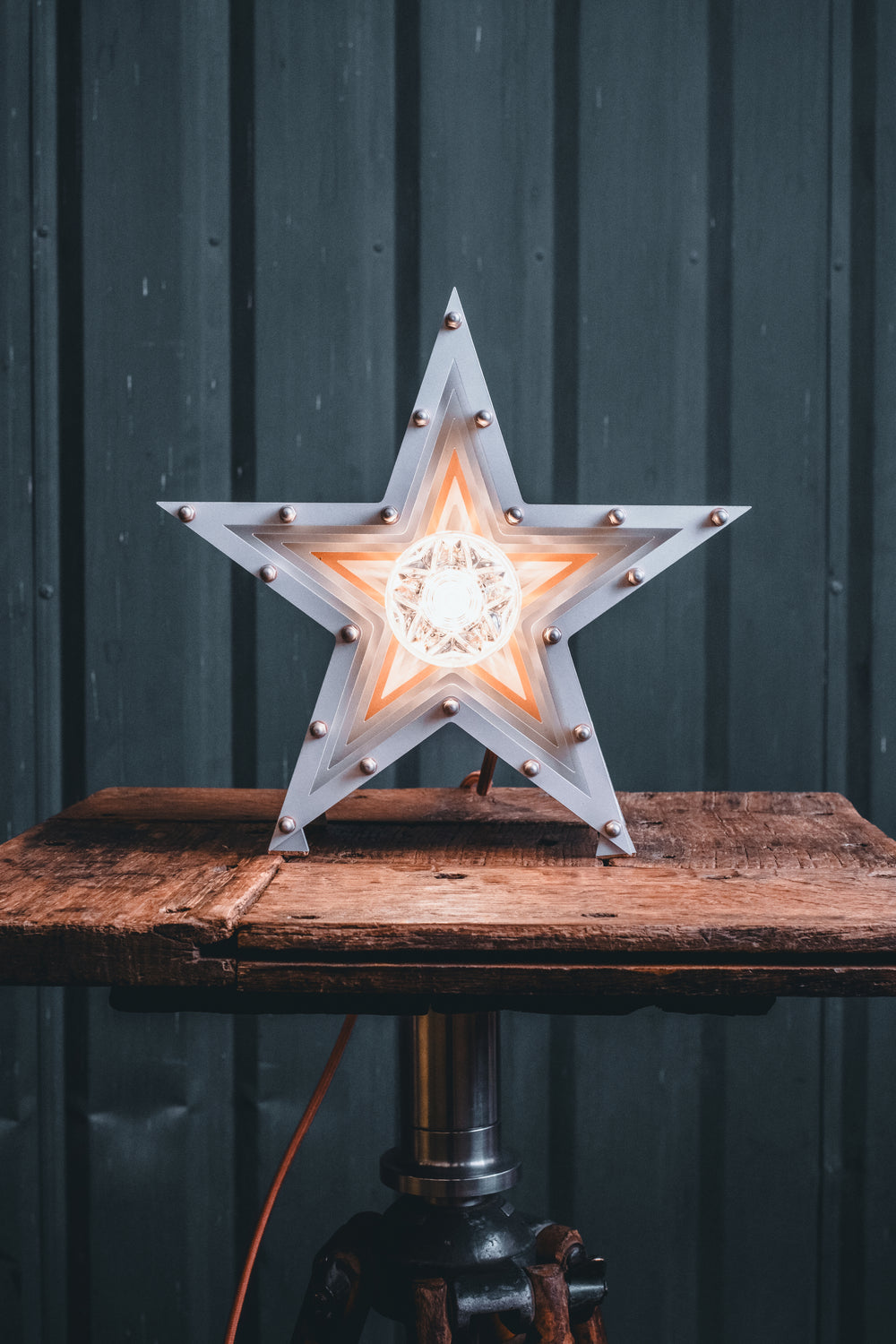 Side view of a Mini Fairground Star lamp in brushed aluminium finish, featuring layered star construction with exposed studding and central illuminated bulb on a rustic wooden table.