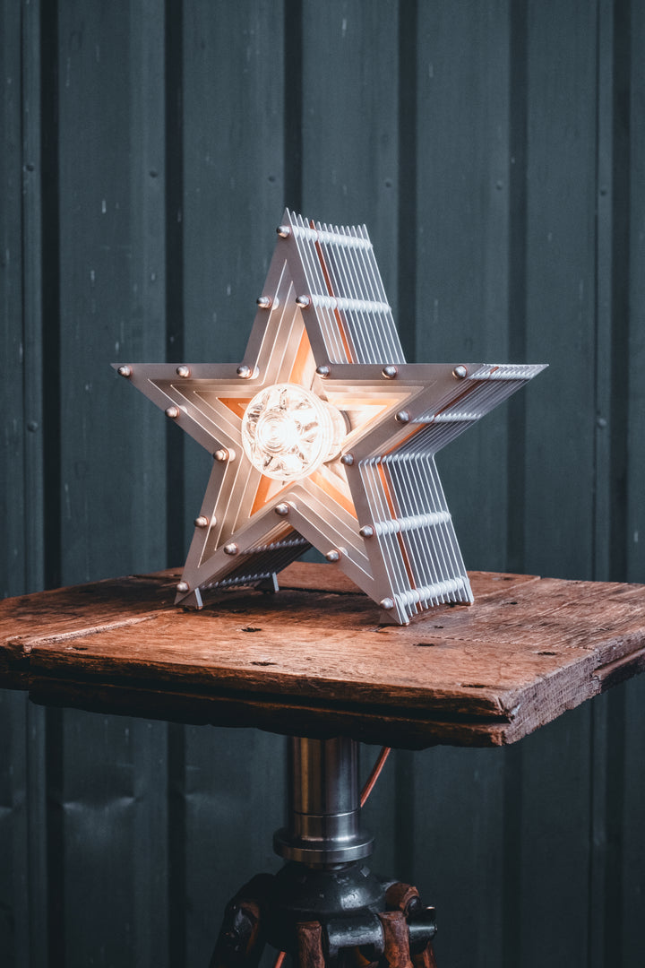 Side view of a Mini Fairground Star lamp in brushed aluminium finish, featuring layered star construction with exposed studding and central illuminated bulb on a rustic wooden table.