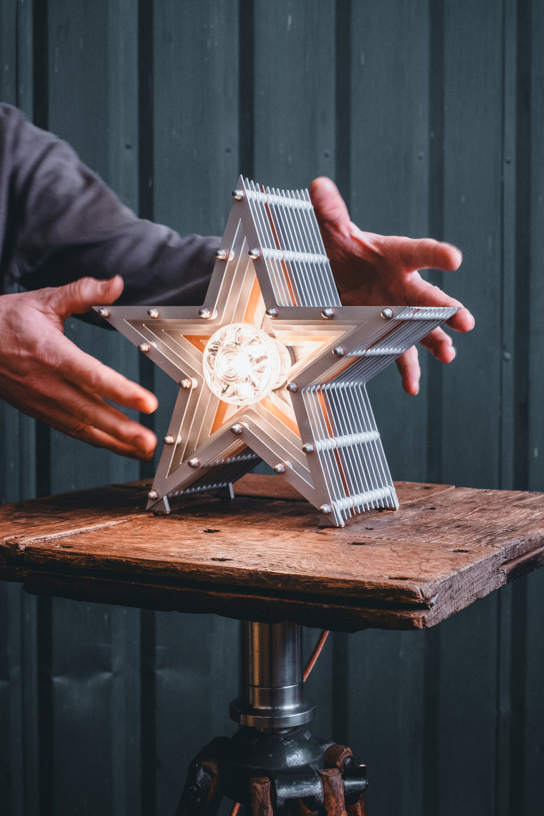 Side view of a Mini Fairground Star lamp in brushed aluminium finish, featuring layered star construction with exposed studding and central illuminated bulb on a rustic wooden table.