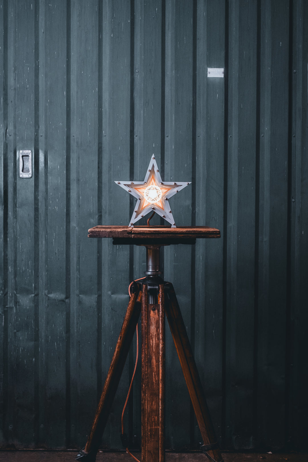 Side view of a Mini Fairground Star lamp in brushed aluminium finish, featuring layered star construction with exposed studding and central illuminated bulb on a rustic wooden table.