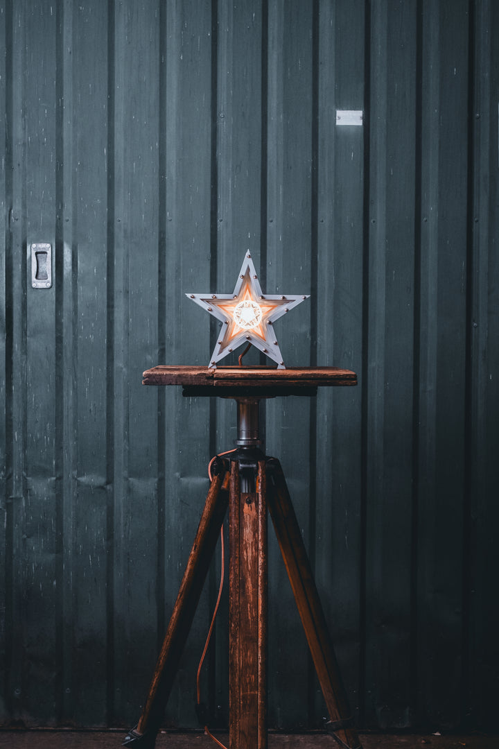 Side view of a Mini Fairground Star lamp in brushed aluminium finish, featuring layered star construction with exposed studding and central illuminated bulb on a rustic wooden table.