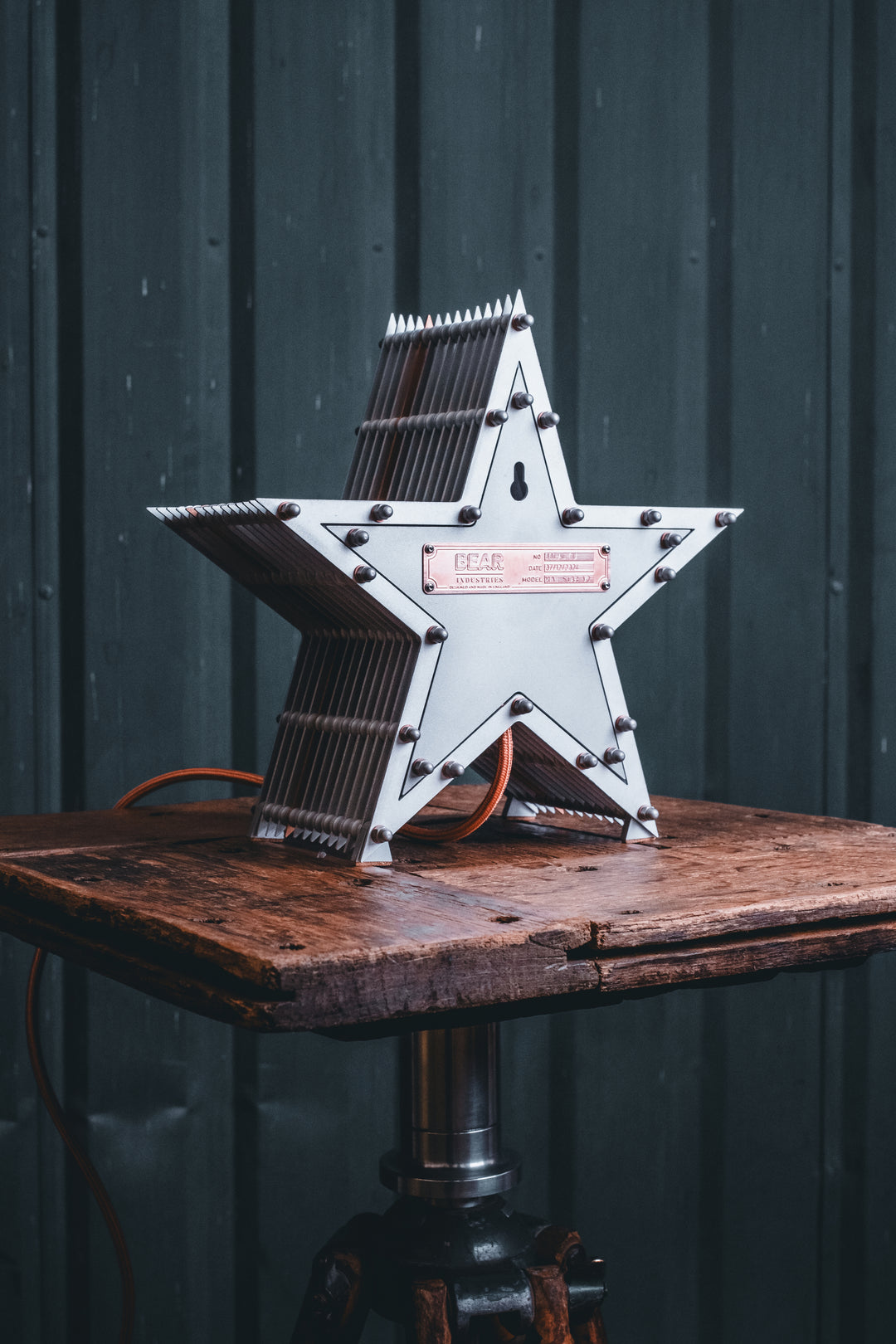 Side view of a Mini Fairground Star lamp in brushed aluminium finish, featuring layered star construction with exposed studding and central illuminated bulb on a rustic wooden table.