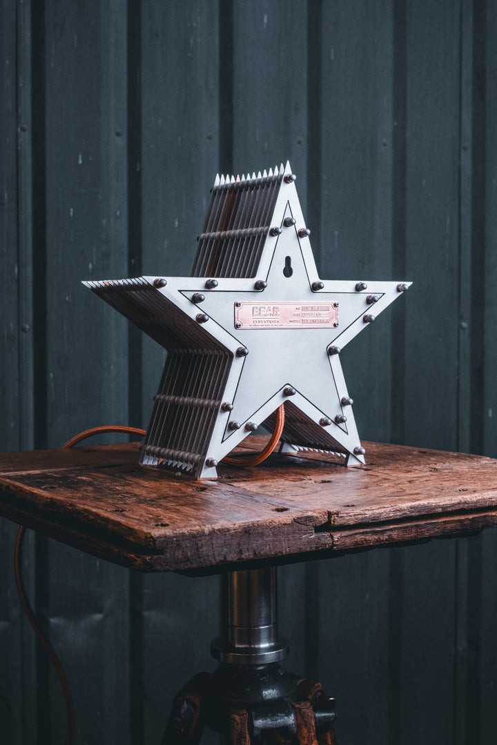 Side view of a Mini Fairground Star lamp in brushed aluminium finish, featuring layered star construction with exposed studding and central illuminated bulb on a rustic wooden table.