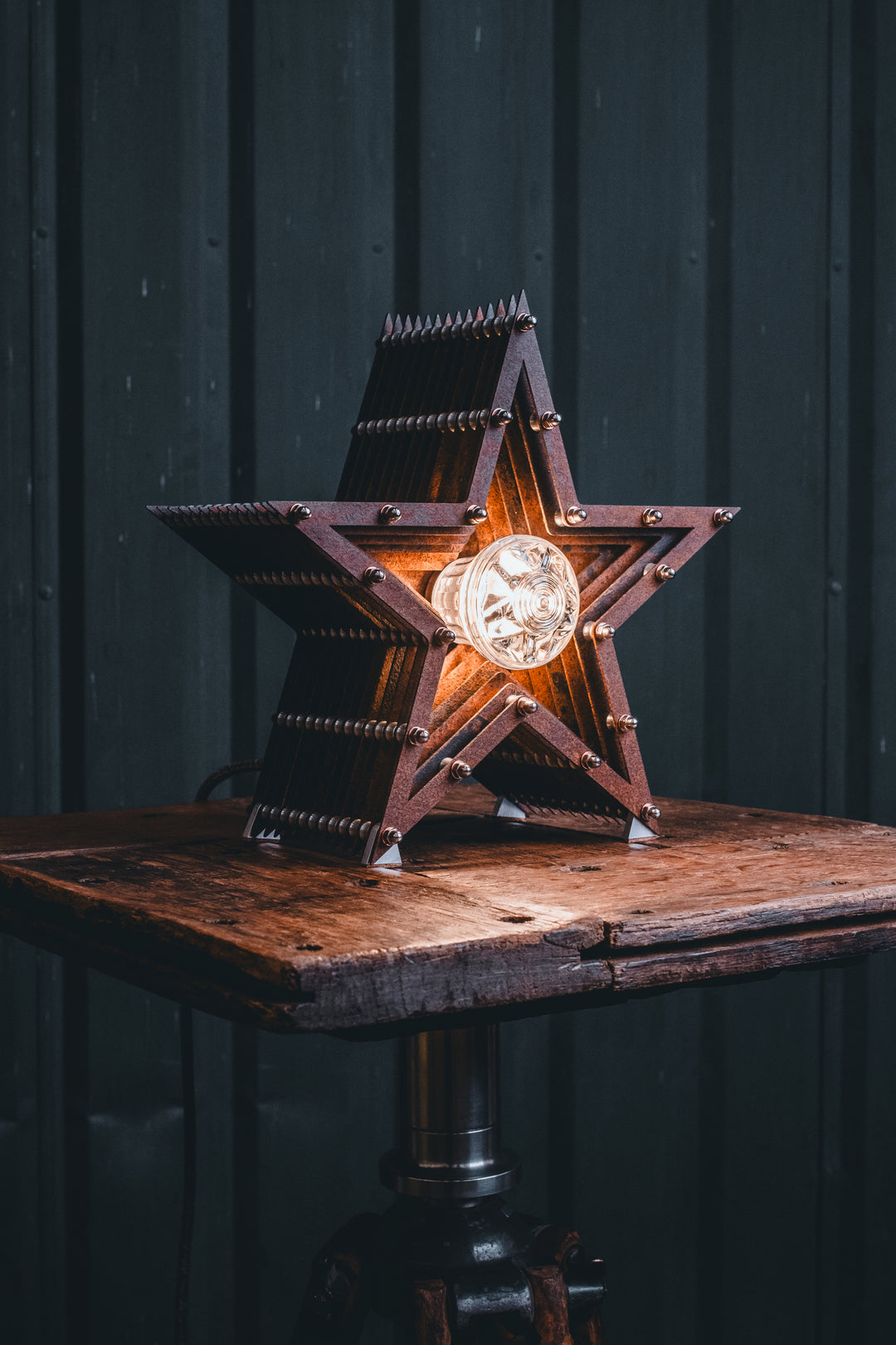 Mini Fairground Star lamp in rust-patinated steel with layered star construction, precision aluminium detailing, and central illuminated bulb, shown on a vintage tripod table.