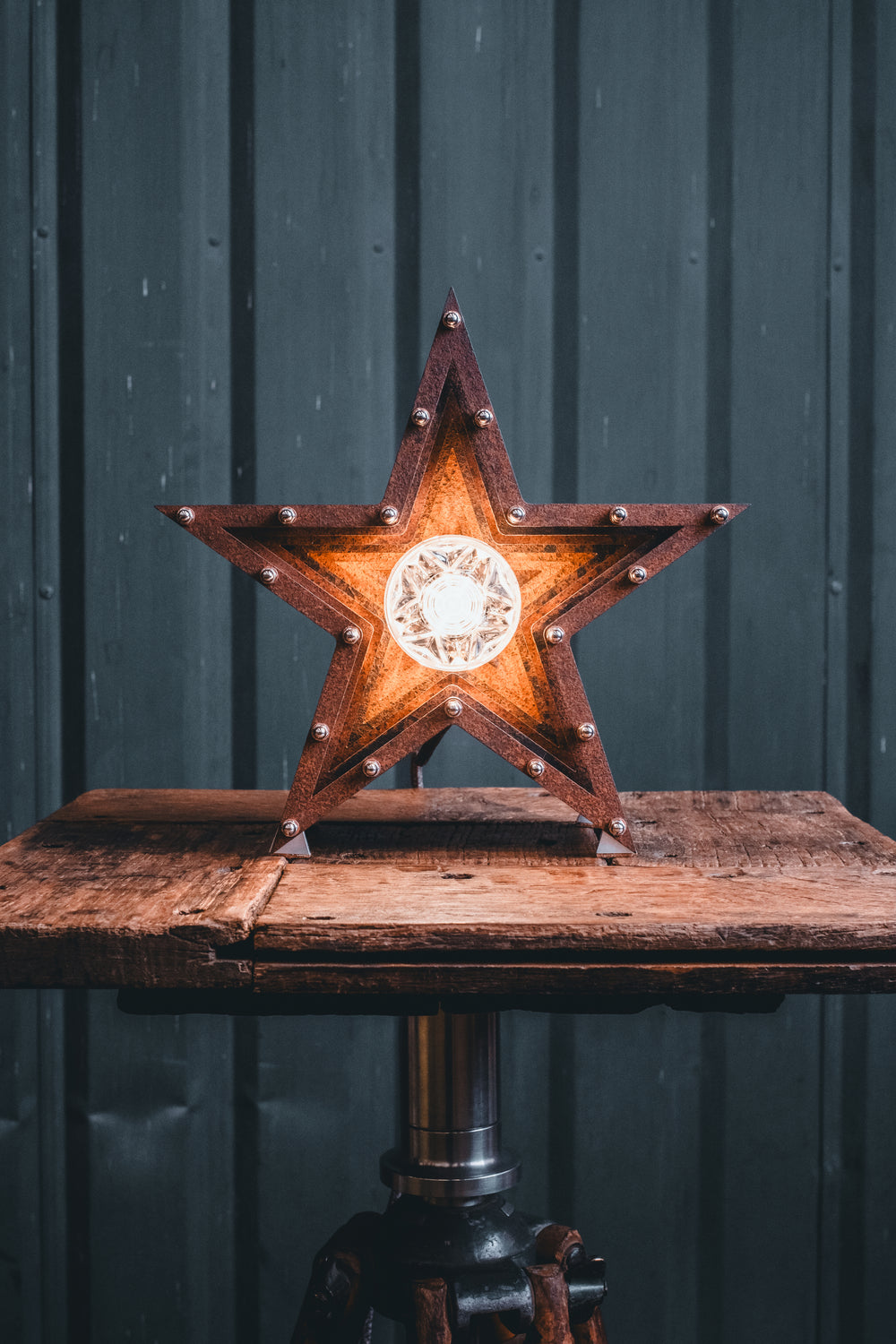 Mini Fairground Star lamp in rust-patinated steel with layered star construction, precision aluminium detailing, and central illuminated bulb, shown on a vintage tripod table.