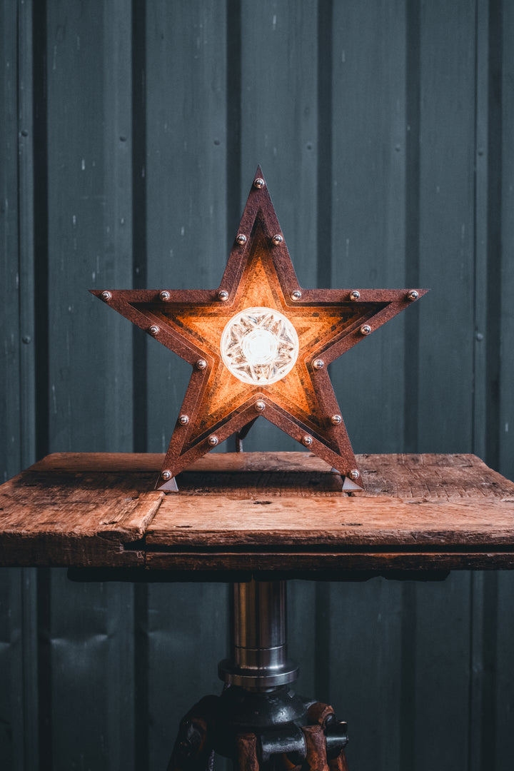 Mini Fairground Star lamp in rust-patinated steel with layered star construction, precision aluminium detailing, and central illuminated bulb, shown on a vintage tripod table.