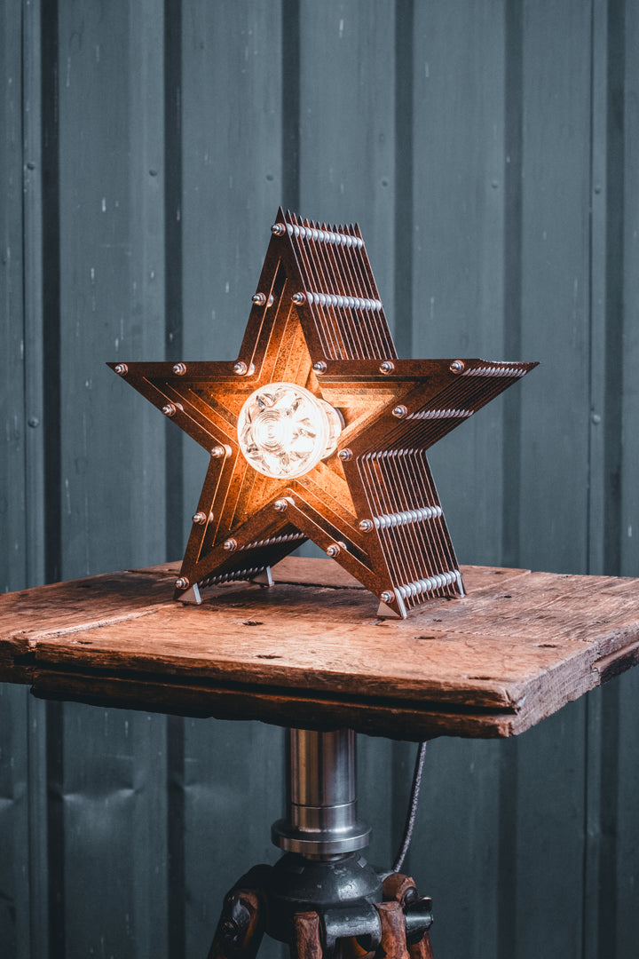 Mini Fairground Star lamp in rust-patinated steel with layered star construction, precision aluminium detailing, and central illuminated bulb, shown on a vintage tripod table.