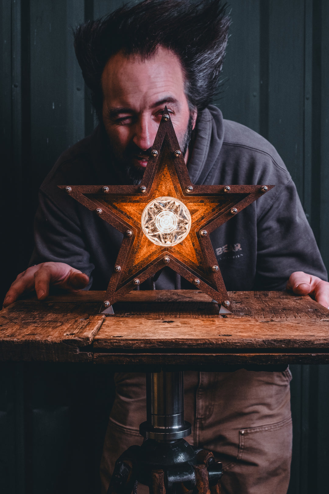 Mini Fairground Star lamp in rust-patinated steel with layered star construction, precision aluminium detailing, and central illuminated bulb, shown on a vintage tripod table.