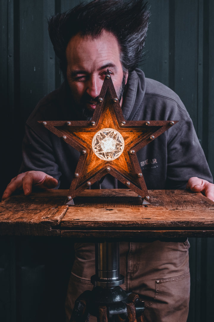 Mini Fairground Star lamp in rust-patinated steel with layered star construction, precision aluminium detailing, and central illuminated bulb, shown on a vintage tripod table.