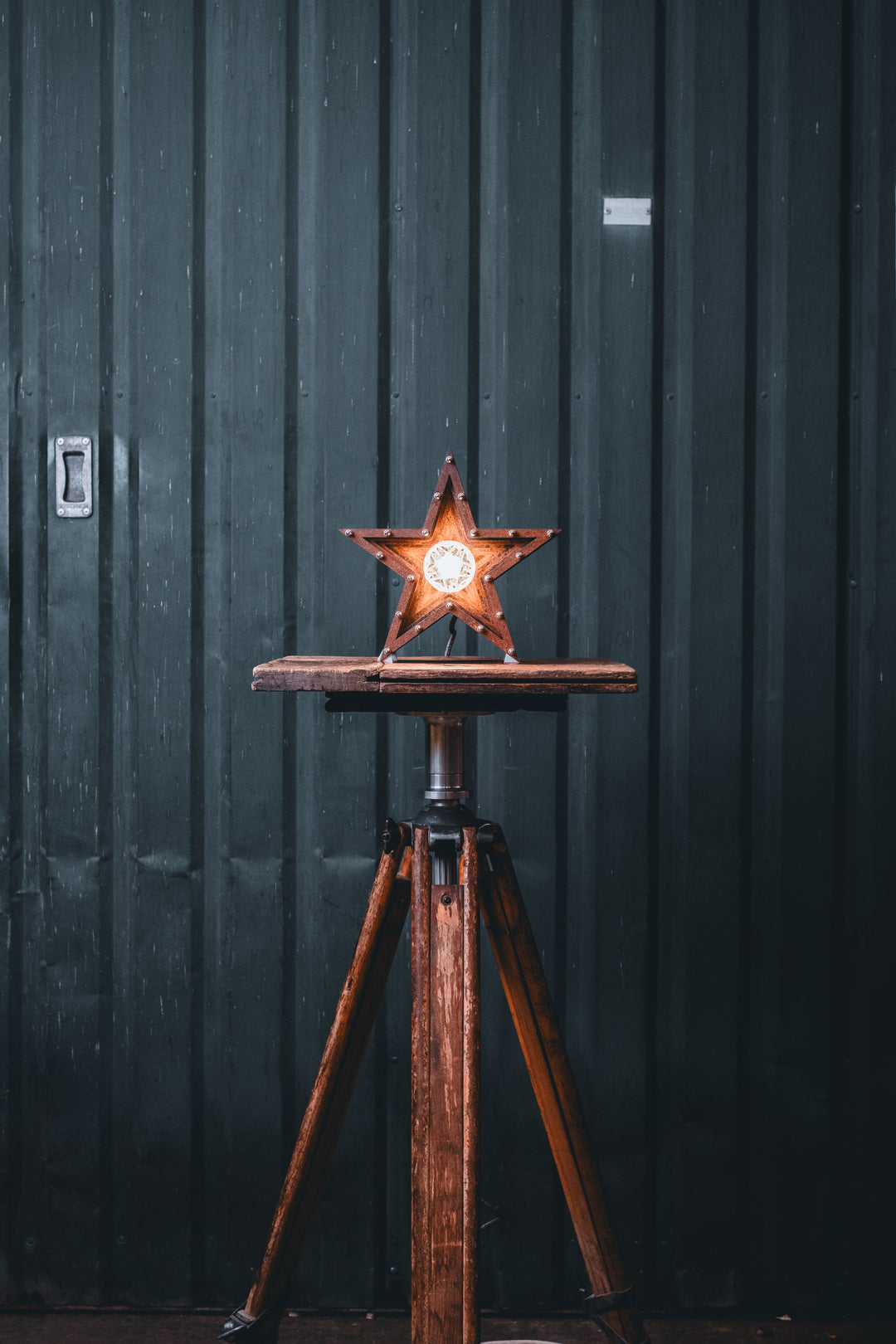 Mini Fairground Star lamp in rust-patinated steel with layered star construction, precision aluminium detailing, and central illuminated bulb, shown on a vintage tripod table.