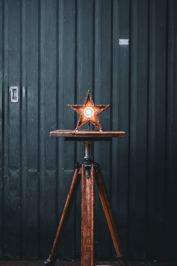 Mini Fairground Star lamp in rust-patinated steel with layered star construction, precision aluminium detailing, and central illuminated bulb, shown on a vintage tripod table.