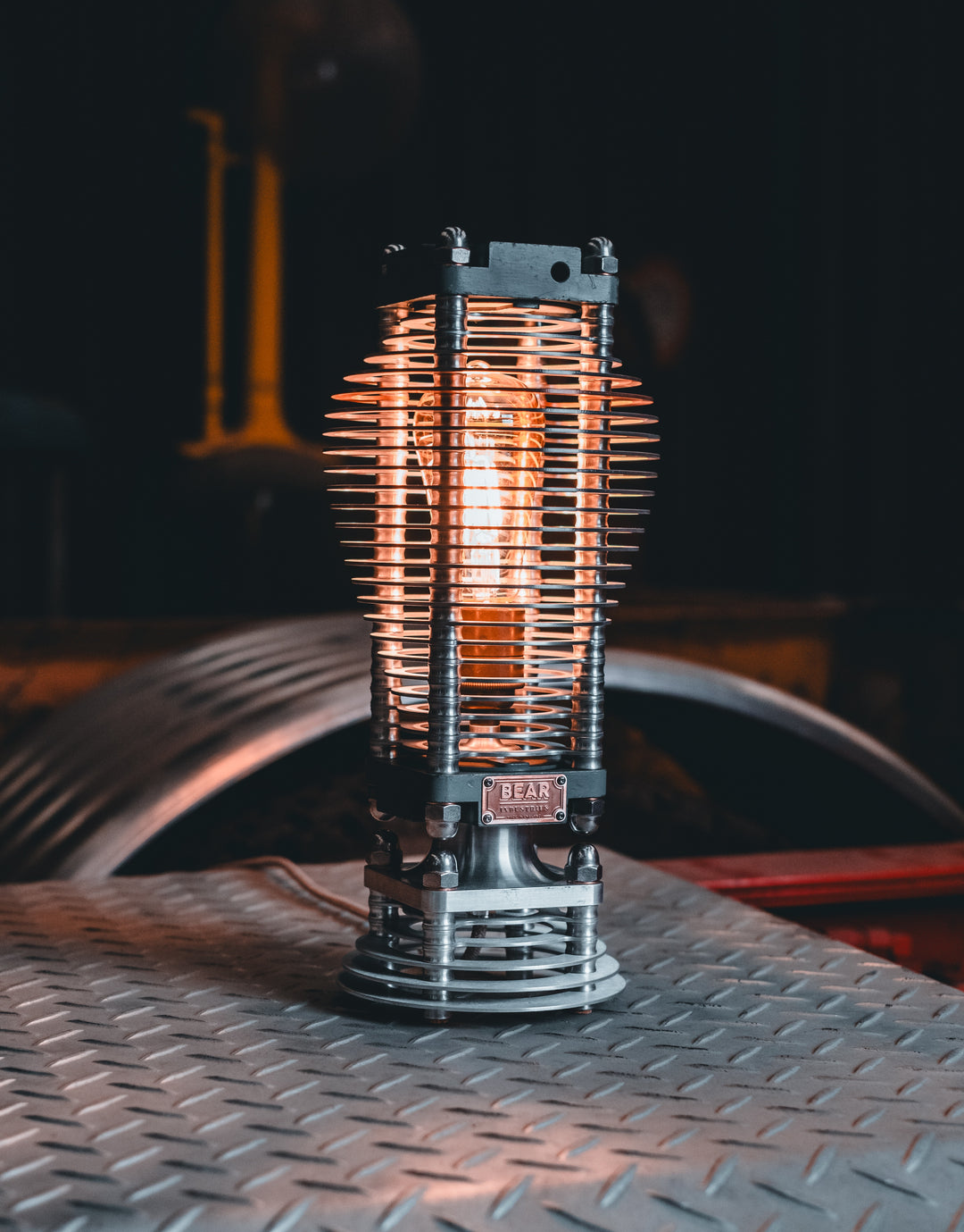 Industrial-style PNEU LAMP handcrafted from disused pneumatic cylinders, featuring layered aluminium and copper fins with a central exposed filament bulb. Designed by Bear Industries as a sculptural lighting piece with a nod to vintage ‘Tilly’ lamps.