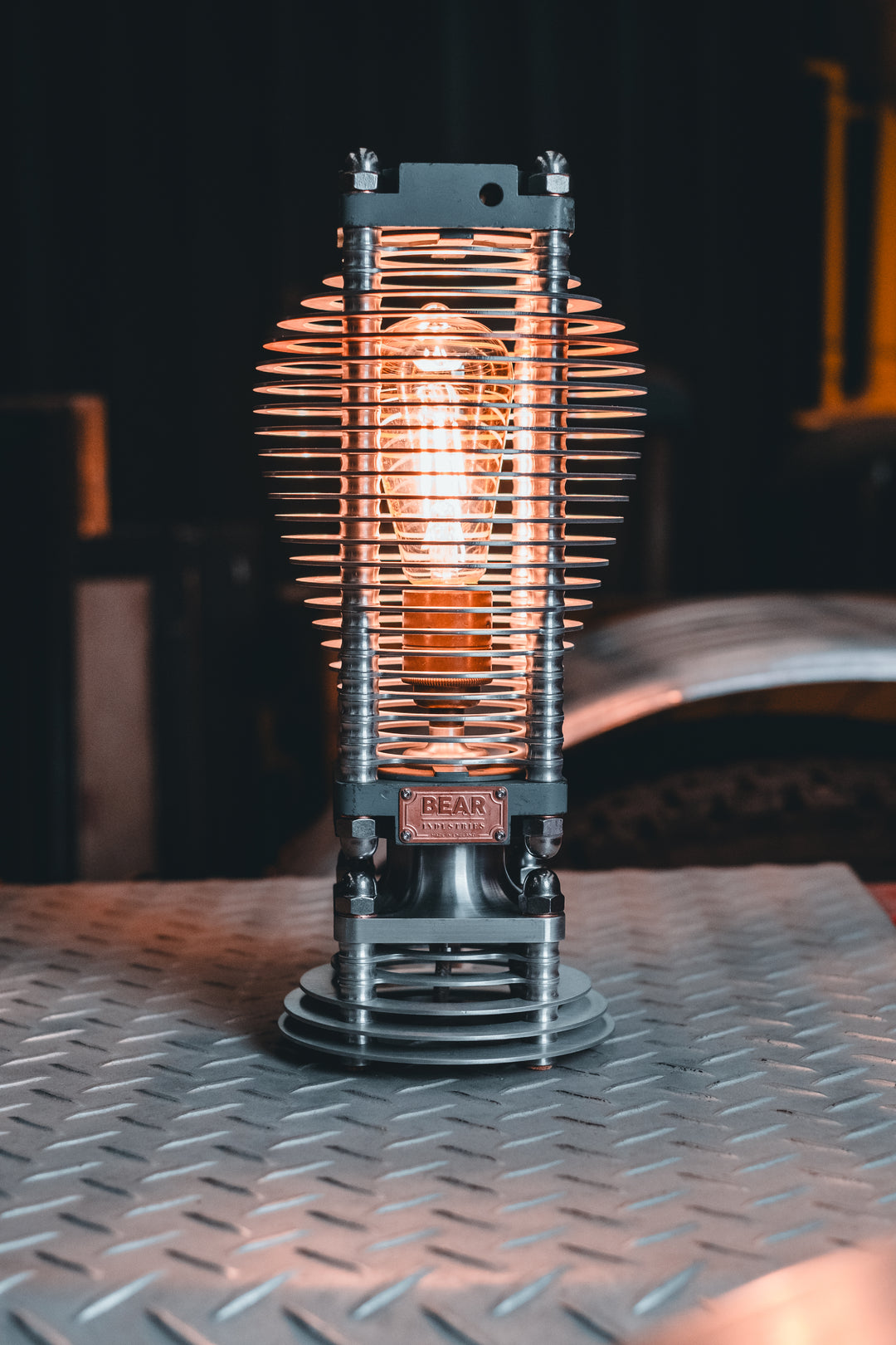 Industrial-style PNEU LAMP handcrafted from disused pneumatic cylinders, featuring layered aluminium and copper fins with a central exposed filament bulb. Designed by Bear Industries as a sculptural lighting piece with a nod to vintage ‘Tilly’ lamps.