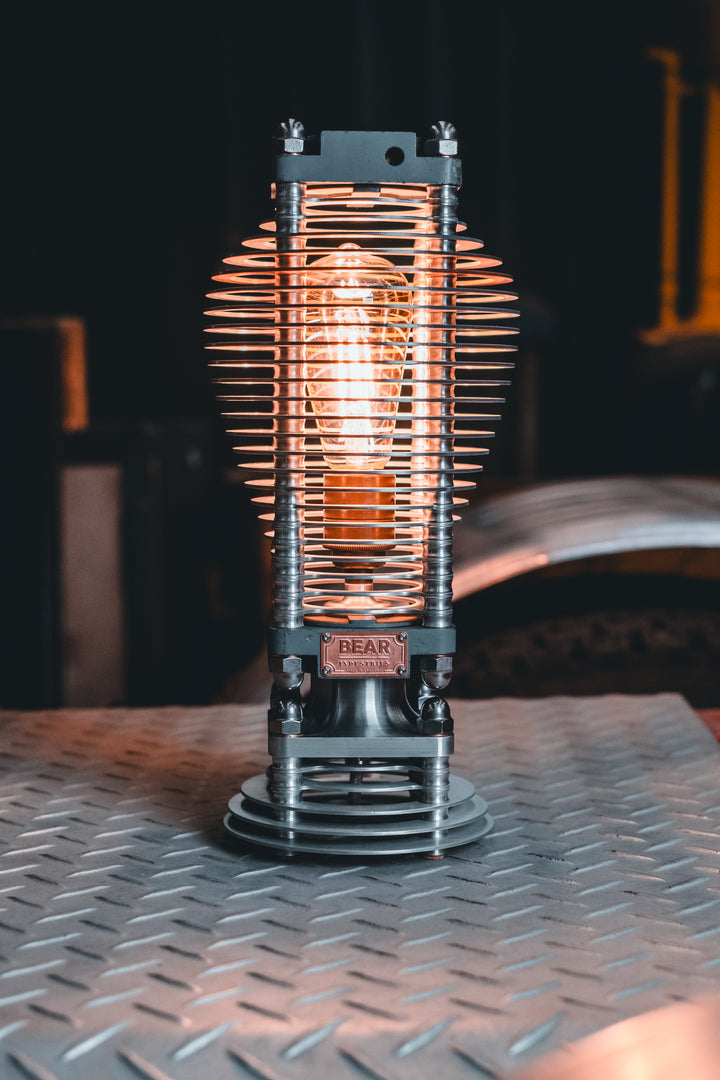 Industrial-style PNEU LAMP handcrafted from disused pneumatic cylinders, featuring layered aluminium and copper fins with a central exposed filament bulb. Designed by Bear Industries as a sculptural lighting piece with a nod to vintage ‘Tilly’ lamps.