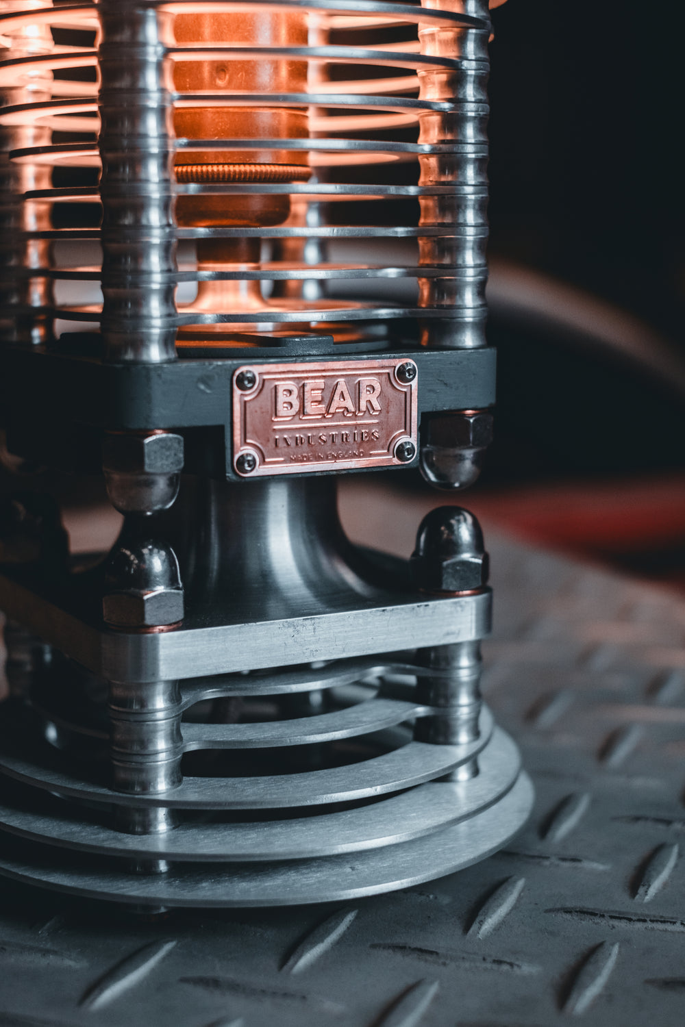 Industrial-style PNEU LAMP handcrafted from disused pneumatic cylinders, featuring layered aluminium and copper fins with a central exposed filament bulb. Designed by Bear Industries as a sculptural lighting piece with a nod to vintage ‘Tilly’ lamps.