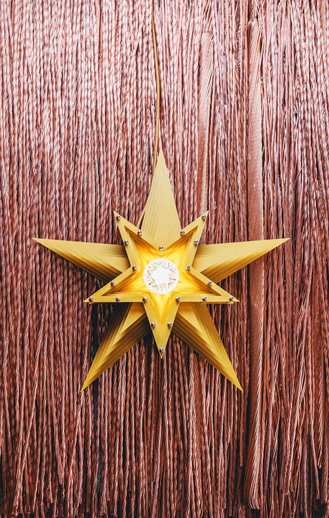 Mini Fairground Sun Star lamp handcrafted from layered steel and aluminium, finished in vibrant yellow with a central lightbulb and mounted against copper wire strands.