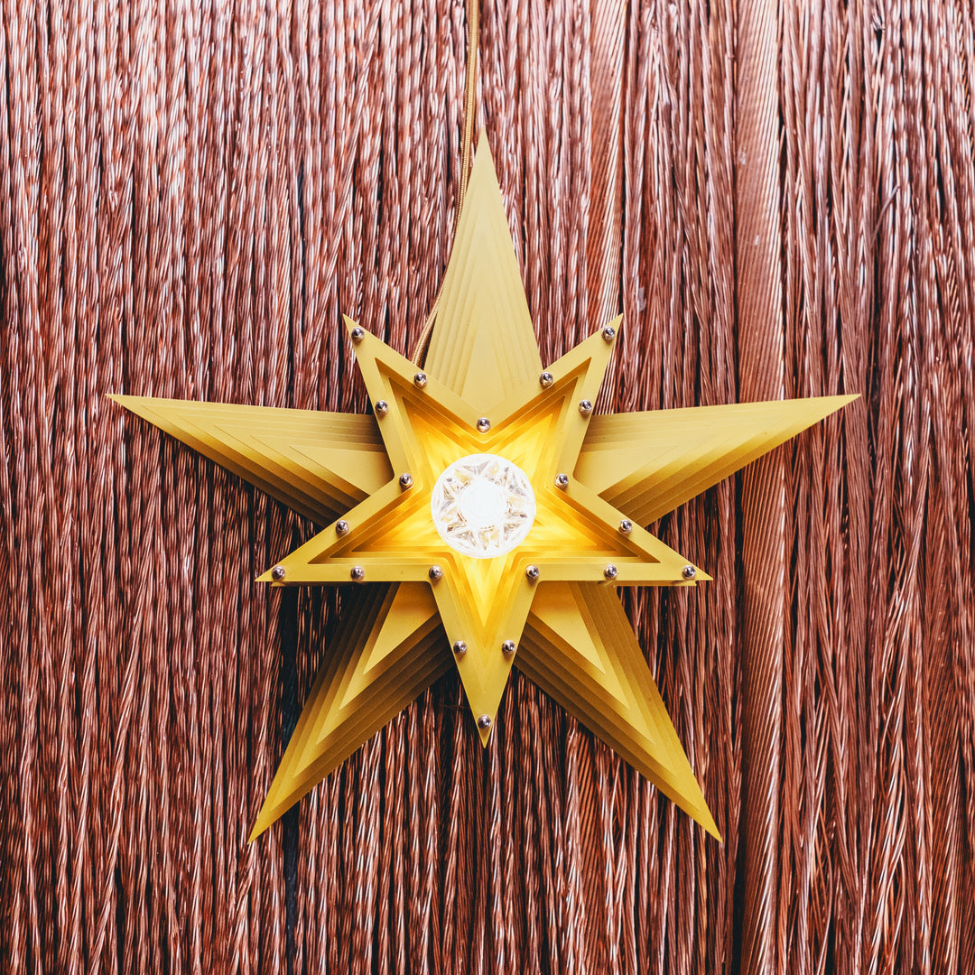 Mini Fairground Sun Star lamp handcrafted from layered steel and aluminium, finished in vibrant yellow with a central lightbulb and mounted against copper wire strands.