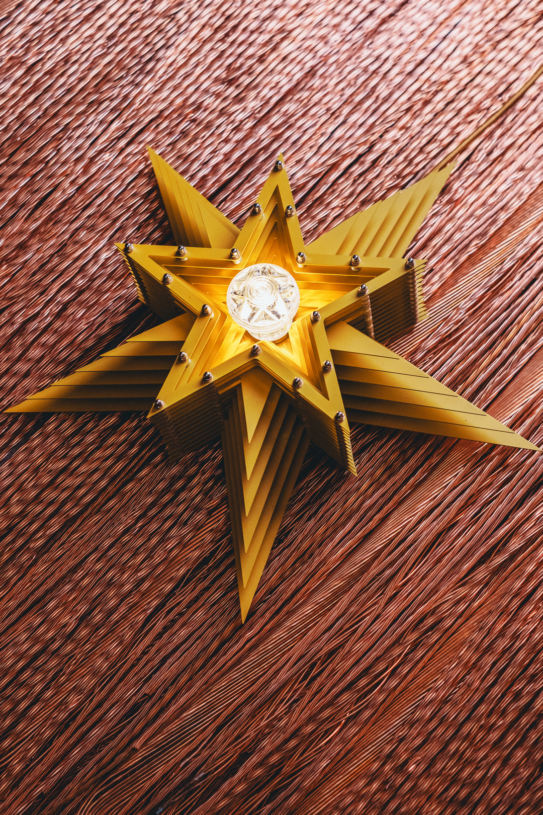 Mini Fairground Sun Star lamp handcrafted from layered steel and aluminium, finished in vibrant yellow with a central lightbulb and mounted against copper wire strands.