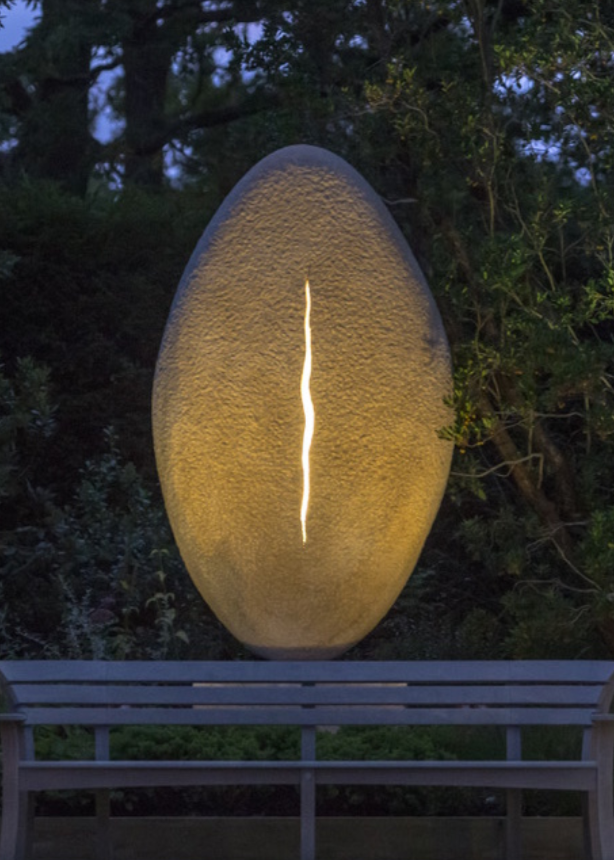 ‘New Beginnings’ by Nicolas Moreton MRSS, a unique commissioned contemporary abstract stone sculpture carved from Ancaster Weatherbed Limestone, featuring a tall, upright oval form with a textured surface and a narrow, irregular vertical opening illuminated by warm white LED light. The sculpture stands on a Portland limestone base in a landscaped garden at dusk, with its illuminated centre contrasting against the dark surrounding trees, purple garden lighting and evening sky.