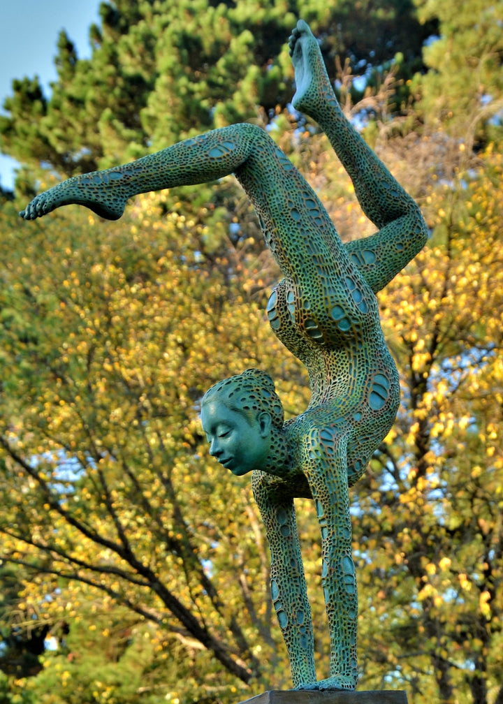Bronze sculpture of a contorted female figure balanced in an acrobatic inverted pose, her body entirely covered in an intricate cellular pattern inspired by plant structures seen under a microscope. Painted in shades of aqua, teal, moss green and warm ochre, the sculpture feels alive with organic energy and movement. Set outdoors against soft greenery, the piece captures the idea of growth, vitality and the hidden structures of nature.