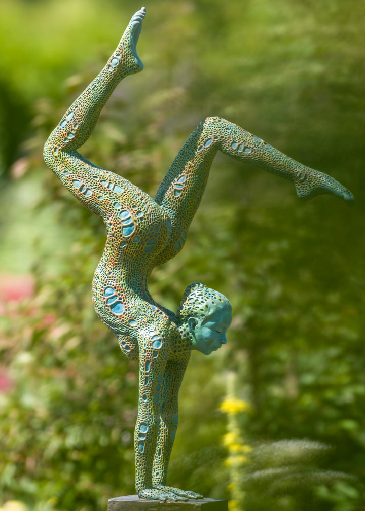 Bronze sculpture of a contorted female figure balanced in an acrobatic inverted pose, her body entirely covered in an intricate cellular pattern inspired by plant structures seen under a microscope. Painted in shades of aqua, teal, moss green and warm ochre, the sculpture feels alive with organic energy and movement. Set outdoors against soft greenery, the piece captures the idea of growth, vitality and the hidden structures of nature.