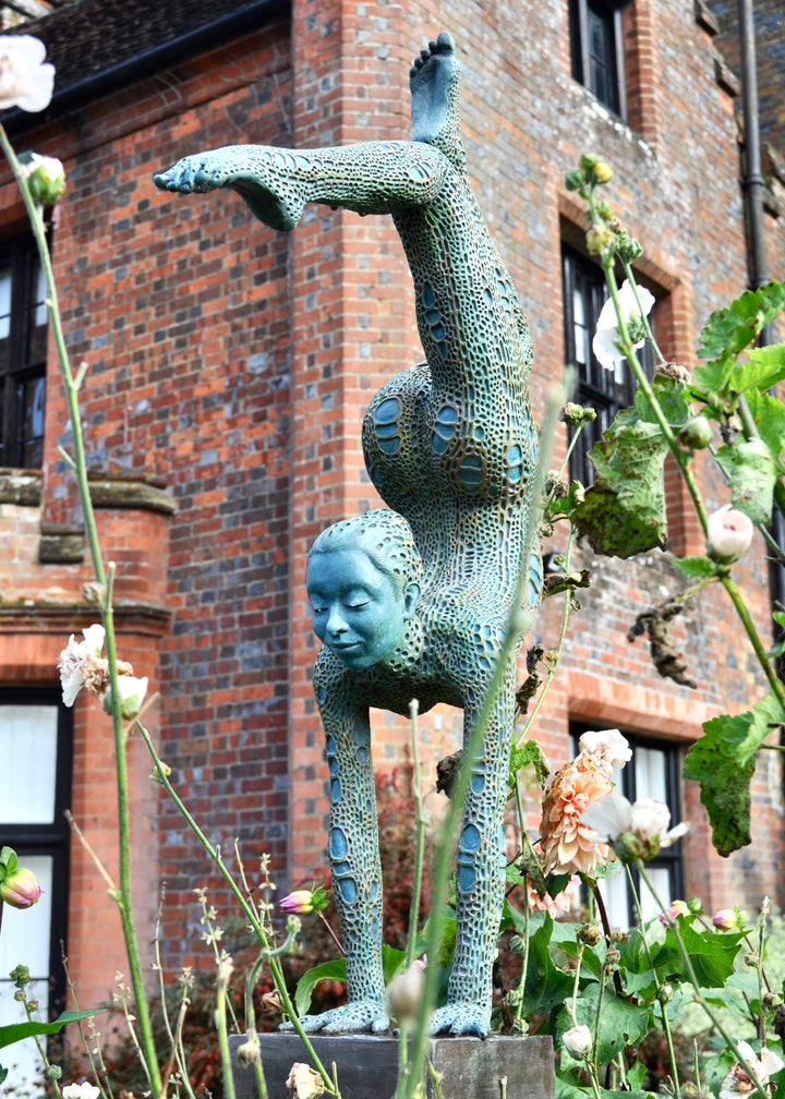 Bronze sculpture of a contorted female figure balanced in an acrobatic inverted pose, her body entirely covered in an intricate cellular pattern inspired by plant structures seen under a microscope. Painted in shades of aqua, teal, moss green and warm ochre, the sculpture feels alive with organic energy and movement. Set outdoors against soft greenery, the piece captures the idea of growth, vitality and the hidden structures of nature.