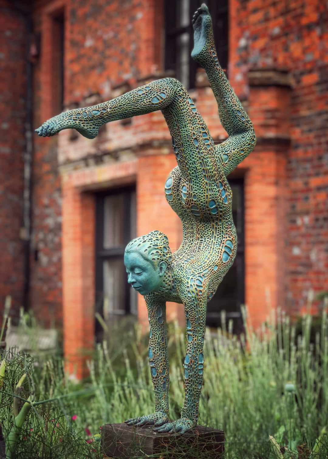 Bronze sculpture of a contorted female figure balanced in an acrobatic inverted pose, her body entirely covered in an intricate cellular pattern inspired by plant structures seen under a microscope. Painted in shades of aqua, teal, moss green and warm ochre, the sculpture feels alive with organic energy and movement. Set outdoors against soft greenery, the piece captures the idea of growth, vitality and the hidden structures of nature.