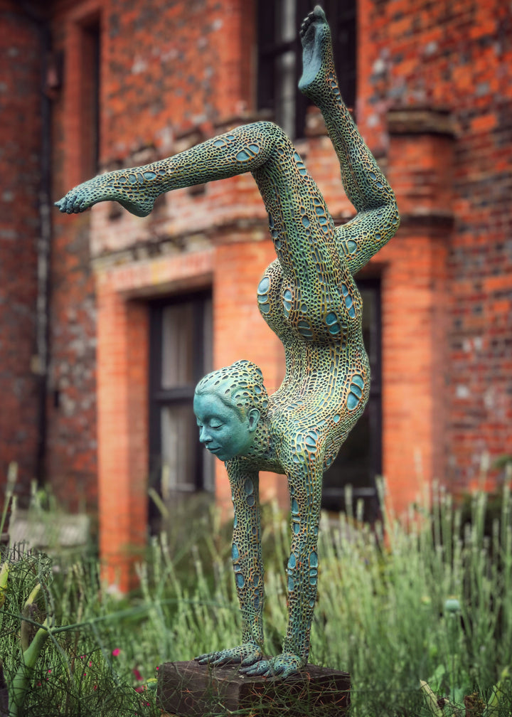 Bronze sculpture of a contorted female figure balanced in an acrobatic inverted pose, her body entirely covered in an intricate cellular pattern inspired by plant structures seen under a microscope. Painted in shades of aqua, teal, moss green and warm ochre, the sculpture feels alive with organic energy and movement. Set outdoors against soft greenery, the piece captures the idea of growth, vitality and the hidden structures of nature.