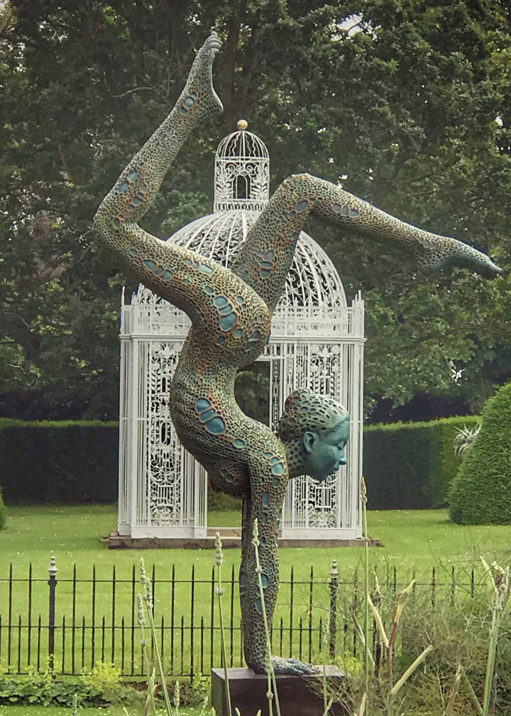 Bronze sculpture of a contorted female figure balanced in an acrobatic inverted pose, her body entirely covered in an intricate cellular pattern inspired by plant structures seen under a microscope. Painted in shades of aqua, teal, moss green and warm ochre, the sculpture feels alive with organic energy and movement. Set outdoors against soft greenery, the piece captures the idea of growth, vitality and the hidden structures of nature.