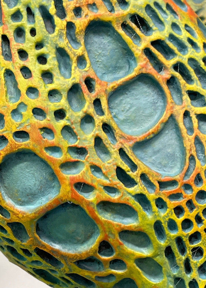 Bronze sculpture of a contorted female figure balanced in an acrobatic inverted pose, her body entirely covered in an intricate cellular pattern inspired by plant structures seen under a microscope. Painted in shades of aqua, teal, moss green and warm ochre, the sculpture feels alive with organic energy and movement. Set outdoors against soft greenery, the piece captures the idea of growth, vitality and the hidden structures of nature.