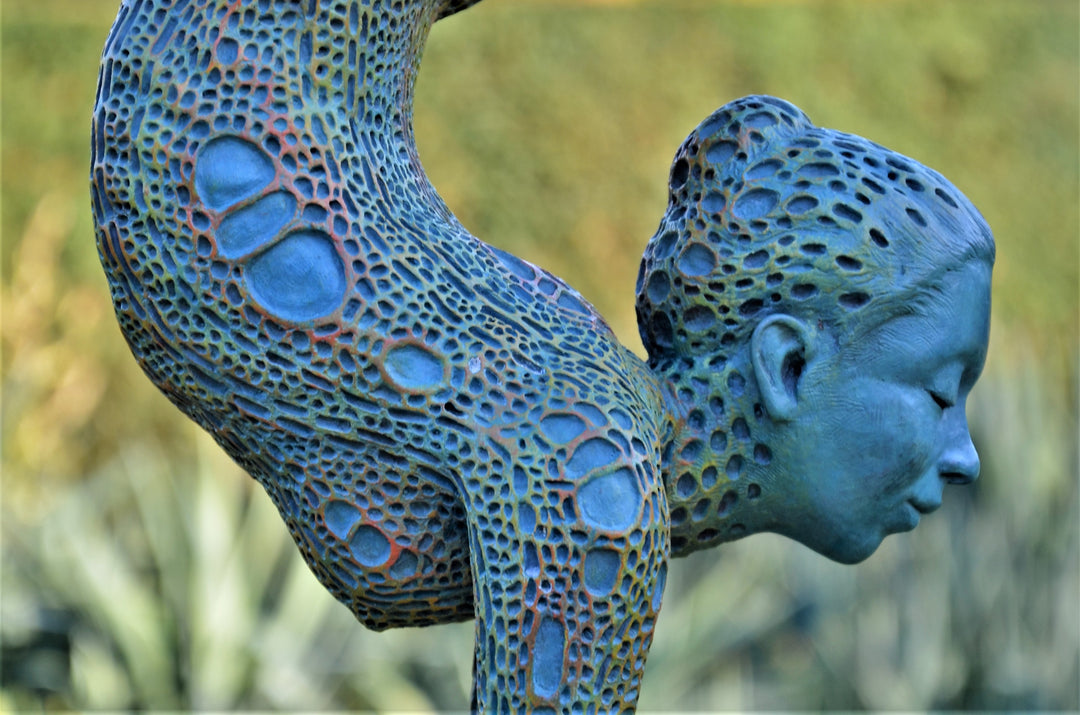 Bronze sculpture of a contorted female figure balanced in an acrobatic inverted pose, her body entirely covered in an intricate cellular pattern inspired by plant structures seen under a microscope. Painted in shades of aqua, teal, moss green and warm ochre, the sculpture feels alive with organic energy and movement. Set outdoors against soft greenery, the piece captures the idea of growth, vitality and the hidden structures of nature.