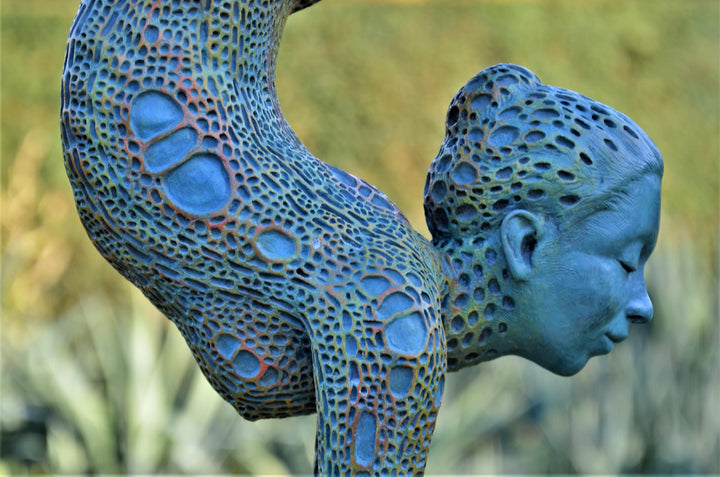 Bronze sculpture of a contorted female figure balanced in an acrobatic inverted pose, her body entirely covered in an intricate cellular pattern inspired by plant structures seen under a microscope. Painted in shades of aqua, teal, moss green and warm ochre, the sculpture feels alive with organic energy and movement. Set outdoors against soft greenery, the piece captures the idea of growth, vitality and the hidden structures of nature.